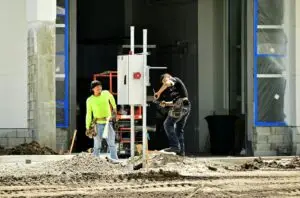 Two construction workers install an electrical panel outside a building site, wearing safety gear.